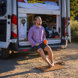 man sitting on bumper of camping van, feet on ocean designs floor mat